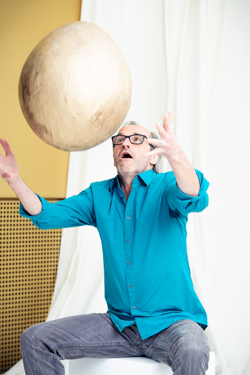 Jürgen Decke, Schauspieler und Geschäftsführer des Theater Pfütze vor goldener Theaterfassade wirft ein riesiges goldenes Ei in die Luft