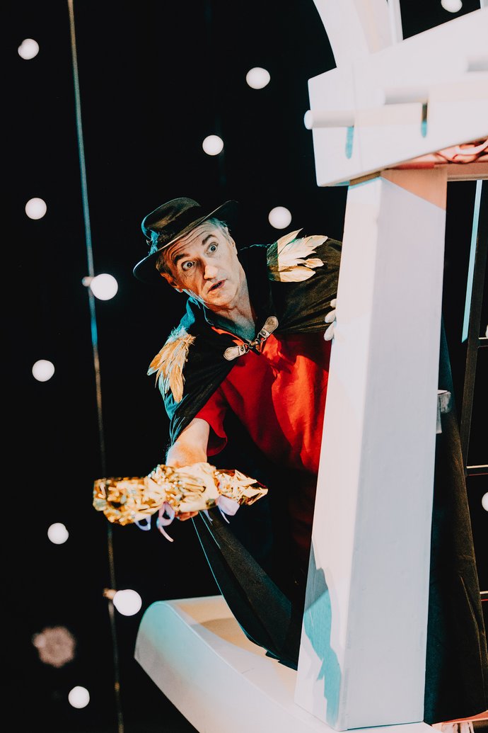 Ein Mann in schwarzem Umhang und mit rotem Hemd hält jemandem ein goldenes, eingepacktes Geschenk hin. Leuchtenden Lichter im dunklen Hintergrund.