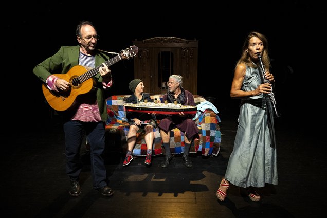 Zwei Musiker:innen stehen vorne am Rand. Einer spielt Gitarre, die andere Flöte. Im Hintergrund sitzen zwei Frauen auf einem Sofa. Die eine wird von der anderen gefüttert.