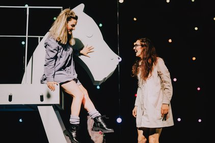 Zwei Frauen auf einer dunkeln Bühne mit Lichtern im Hintergrund, eine sitzt auf einem großen weißen Schachpferd, die andere steht daneben, beide tragen lange Hemdem und Stiefel.