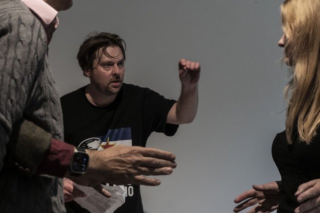 Ein Mann in Jeans und T-Shirt spricht erregt. Zwei Personen, die links und recht am Rande des Fotos stehen, beobachten ihn dabei.
