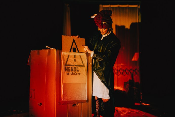 Mann mit roter gestrickter Kopfbedeckung und grünen Mantel hält ein Schild mit einem Warnsymbol und dem Text 'HENDL with Care' vor einem großen offenem Karton hoch.