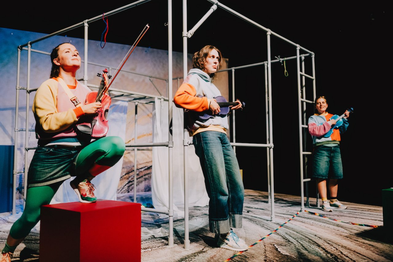 Drei Schauspielerinnen in bunter Kleidungsind nebeneinander an ein Klettergerüst gelehnt. Alle haben bunte Geigen und spielen sie auf ungewöhnliche Weise. Eine Geige wird wie eine Gitarre gehalten.