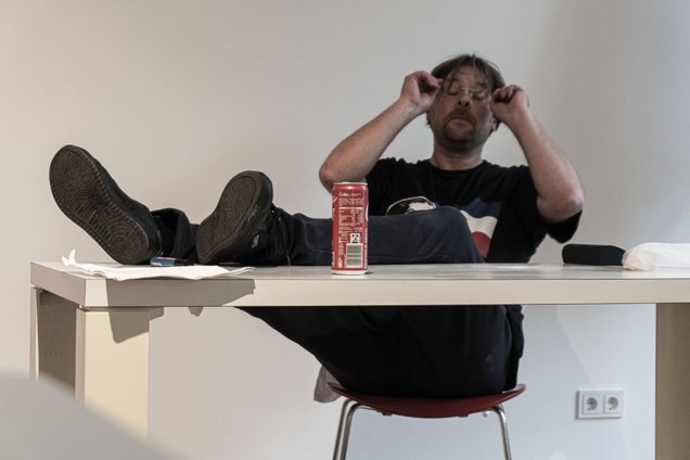 Ein Mann in Jeans und T-Shirt, Mitte vierzig, sitzt auf einem Stuhl und legt die Füße auf einem Tisch vor ihm ab. Er reibt sich die Augen und scheint erschöpft.