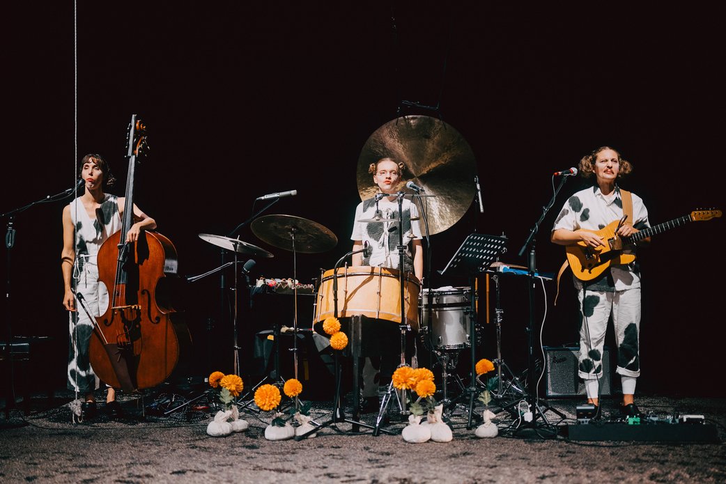 Die Band mit ihren Instrumenten: Links eine Kontrabassistin, mittig ein Schlagzeuger, rechts ein Gitarrist. Alle sind als Kühe verkleidet und kauen. Der Hintergrund ist schwarz. 