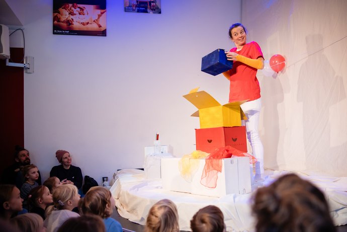 Frau steht auf einer Bühne und hält eine blaue Kiste über einem Stapel bunter Kisten vor einem sitzenden Publikum aus Kindern und Erwachsenen.