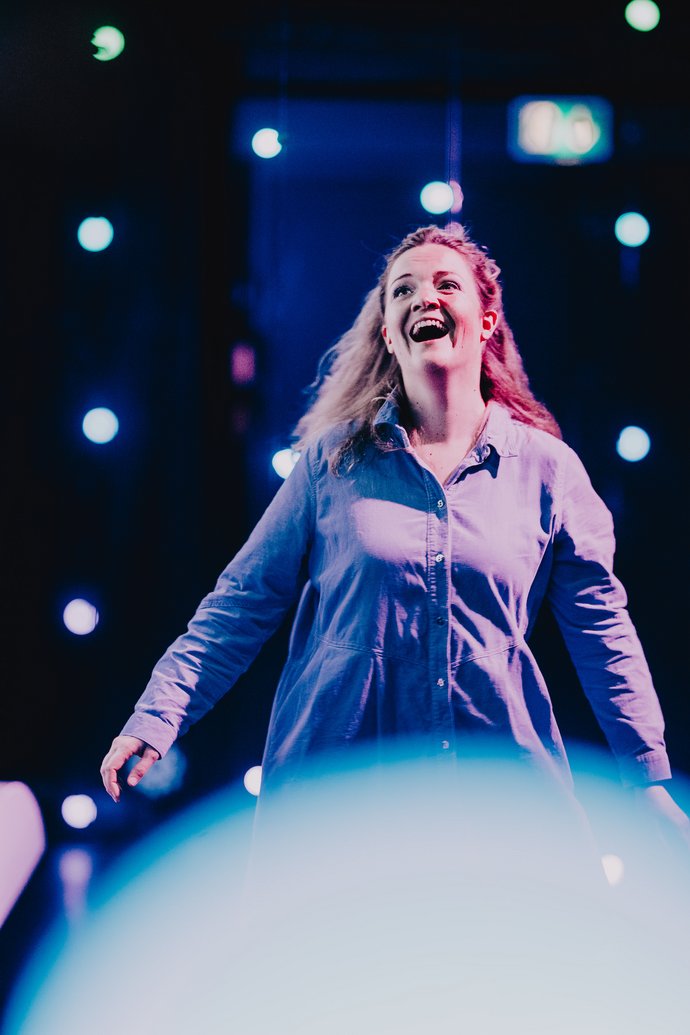 Eine junge Frau mit offenem, langem Haar und lila Cordbluse lacht ausgelassen und in Bewegung. Der Raum ist dunkel mit blau leuchtenden Lichter.