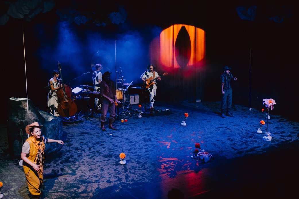 Die ganze Bühne ist in blaues Licht getaucht. Im Hintergrund sieht man die Band (Kontrabassistin, Schlagzeuger, Gitarrist), vorn links eine Schauspielerin in gelbem Cowboykostüm mit geschlossenen Augen. Auf einer Lamellenwand rechts ist ein großes rotes Auge mit schwarzer, schlitzförmiger Pupille projiziert.