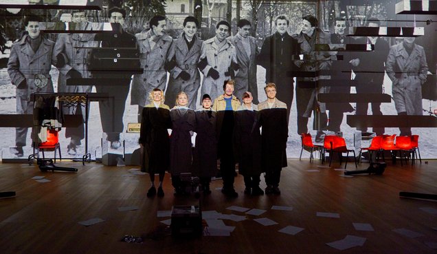 Vier Darsteller:innen, Körper verdeckt durch eine schwarze Decke, schauen ins Publikum. Auf der Wand hinter ihnen sich schwarz-weiß Fotos aus vergangenen Zeiten zu sehen.