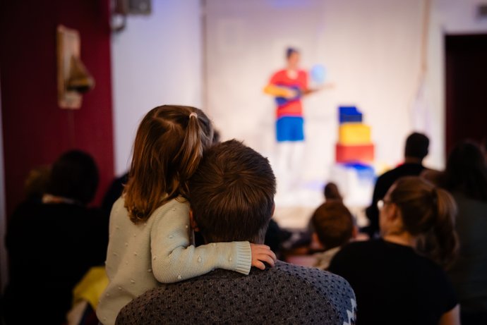 Kind sitzt auf dem Schoß einer erwachsenen Person, beide von hinten fotografiert und schauen auf eine unscharfe Bühne mit einer Person, ein Instrument und bunten Kisten.