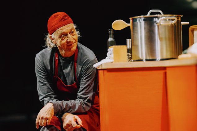 Mann mit roter Kochmütze und roter Schürze sitzt zufrieden mit hochgezogenen Augenbrauen neben einem orangefarbenen Küchentresen mit einem großen Edelstahltopf, einer Flasche und einem Holzlöffel.