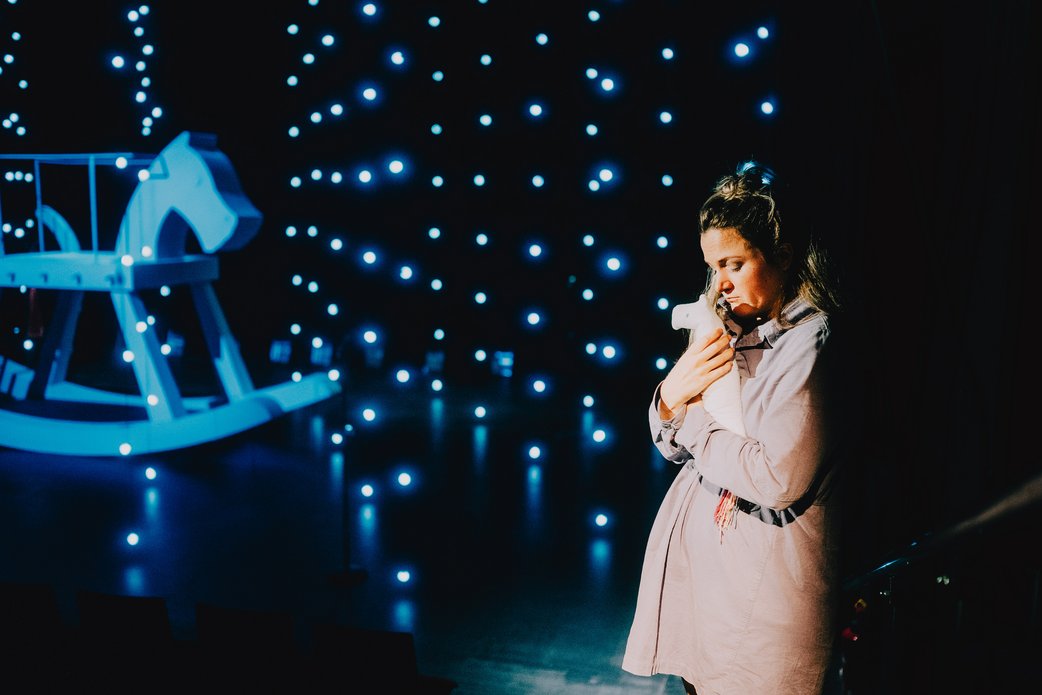 Eine junge Frau in hellem Mantel hält ein Kuscheltier im Arm. Der Bühnenraum ist schwarz, sie ist umgeben von blau leuchtenden Lichtern. Hinten links steht ein riesiges weißes Schaukelpferd.