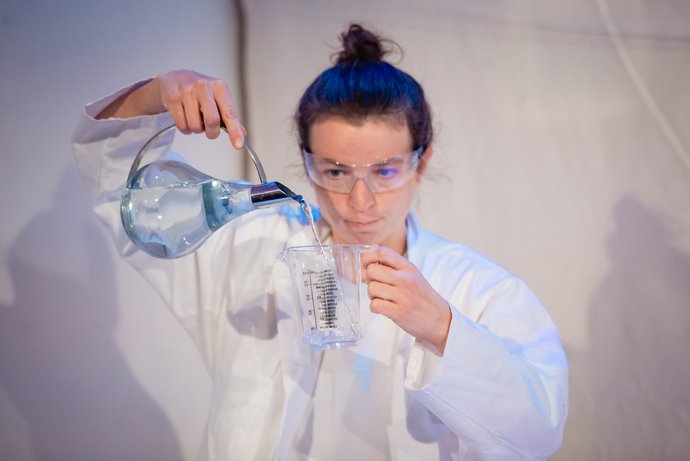 In einem wießem Kittel gießt eine Frau mit Schutzbrille Wasser aus einem Krug in einen Messbecher, mit weißem Hintergrund.