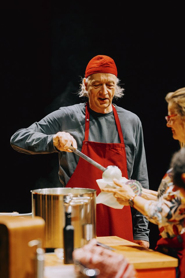 Person mit roter Kochmütze und roter Schürze schöpft Suppe aus einem großen Topf in die Schüssel einer anderen Person.