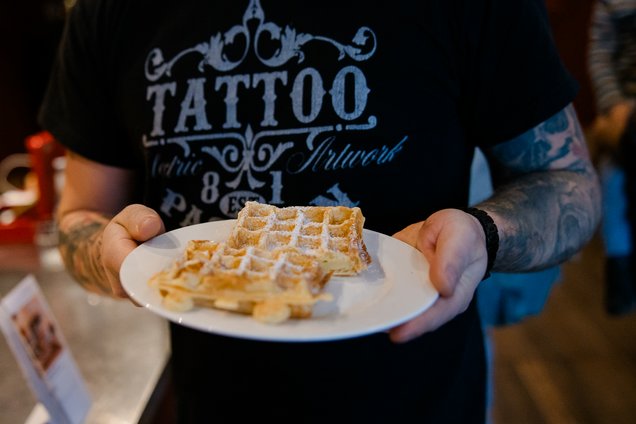 Person mit tätowierten Armen hält einen weißen Teller mit zwei Waffeln, die mit Puderzucker bestreut sind. 