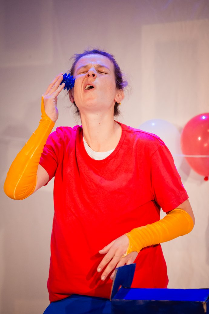 Frau mit rotem T-Shirt und gelben Armstulpen bewegt einen blauen Ball an ihrer rechte Backe, im Hintergrund ist ein rote und weißer Luftballon zu sehen.