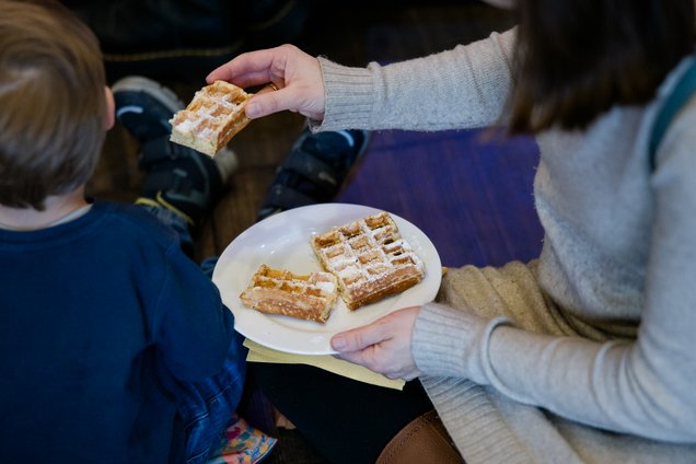Von oben aufgenommenes Bild einer sitzenden Person, die einem Kind ein Stück Waffel von einem Teller mit mehreren Waffelstücken reicht. 