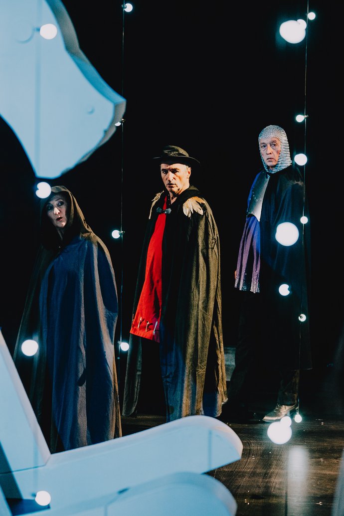 Drei Personen in dunklen Umhängen schauen vor schwarzem Hintergrund neugierig zu uns. Helle Lichterketten hängen im Raum. Im Vordergrund Kopf und Kufen eines großen, weißen Schaukelpferds.