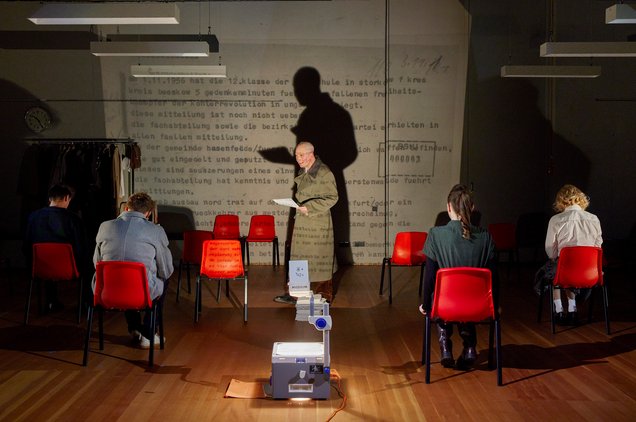 Es steht ein Mann im Mantel auf der Bühne. Er wirft einen großen Schatten an die Wand hinter ihm durch einen Overhead-Projektor, der vor ihm steht. Einige Menschen sitzen auf Stühlen, der Rücken zum Publikum. Rote Stühle stehen verteilt im Raum, eine Neonlampe hängt von oben herab. Im Hintergrund ist ein schwarz-weiß an die Wand projiziert.