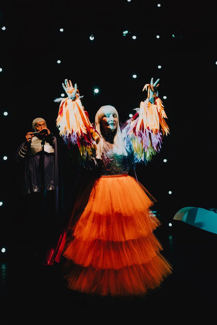 Eine Frau mit weißblondem Haar in einem mehrlagigen, orangefarbenen Rock mit bunten, federbesetzten Oberteil hebt die Arme vor einem dunklen Hintergrund mit kleinen Lichtpunkten. Im Hintergrund spielt ein Mann Mundharmonika.