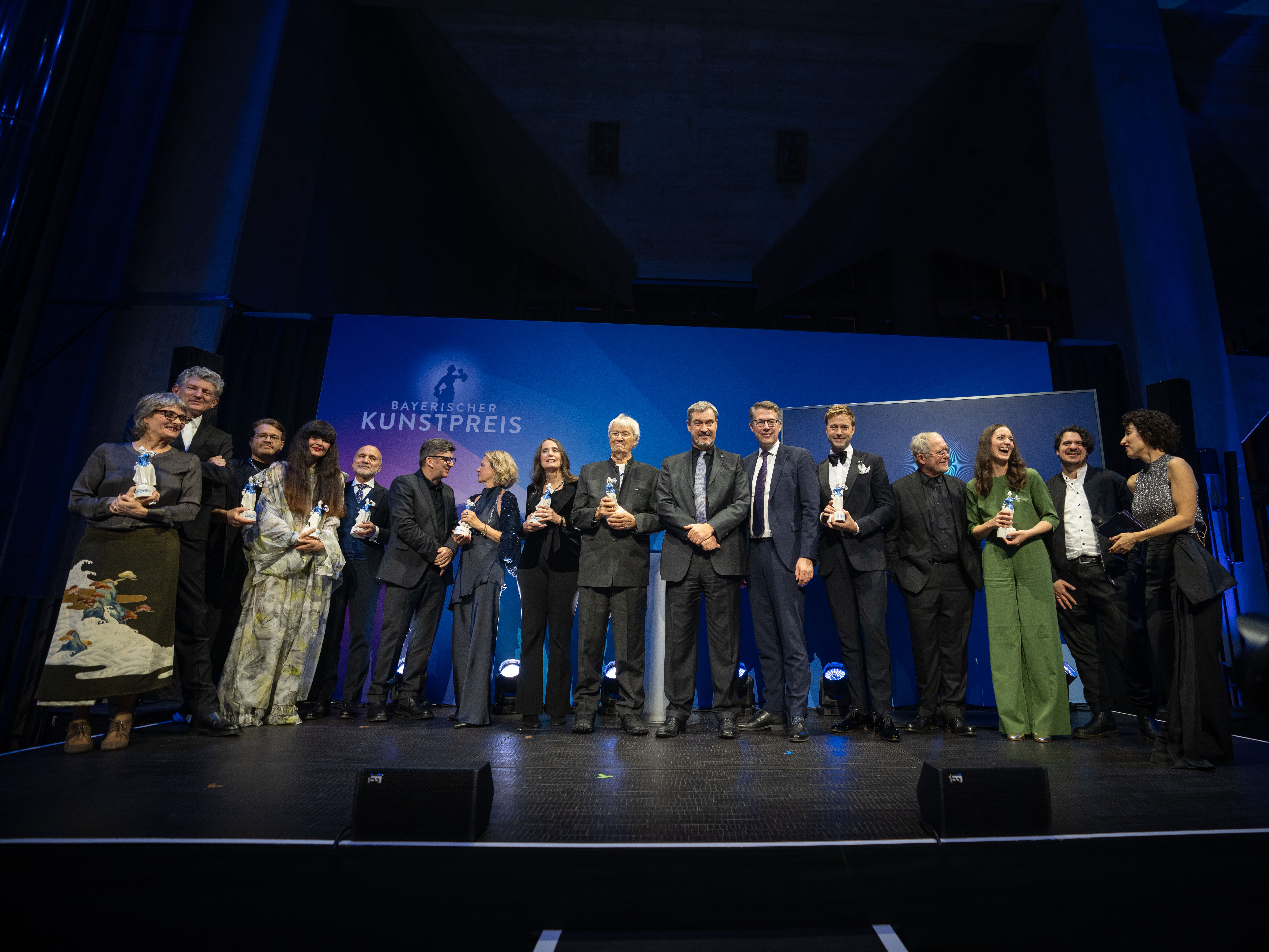 Die Preisträger·innen des Bayerischen Kunstpreises in allen Kategorien sind auf der Bühne vor blauem Hntergrund versammelt. In der Mitte stehen Gerhard Polt, Markus Söder und Markus Blume.