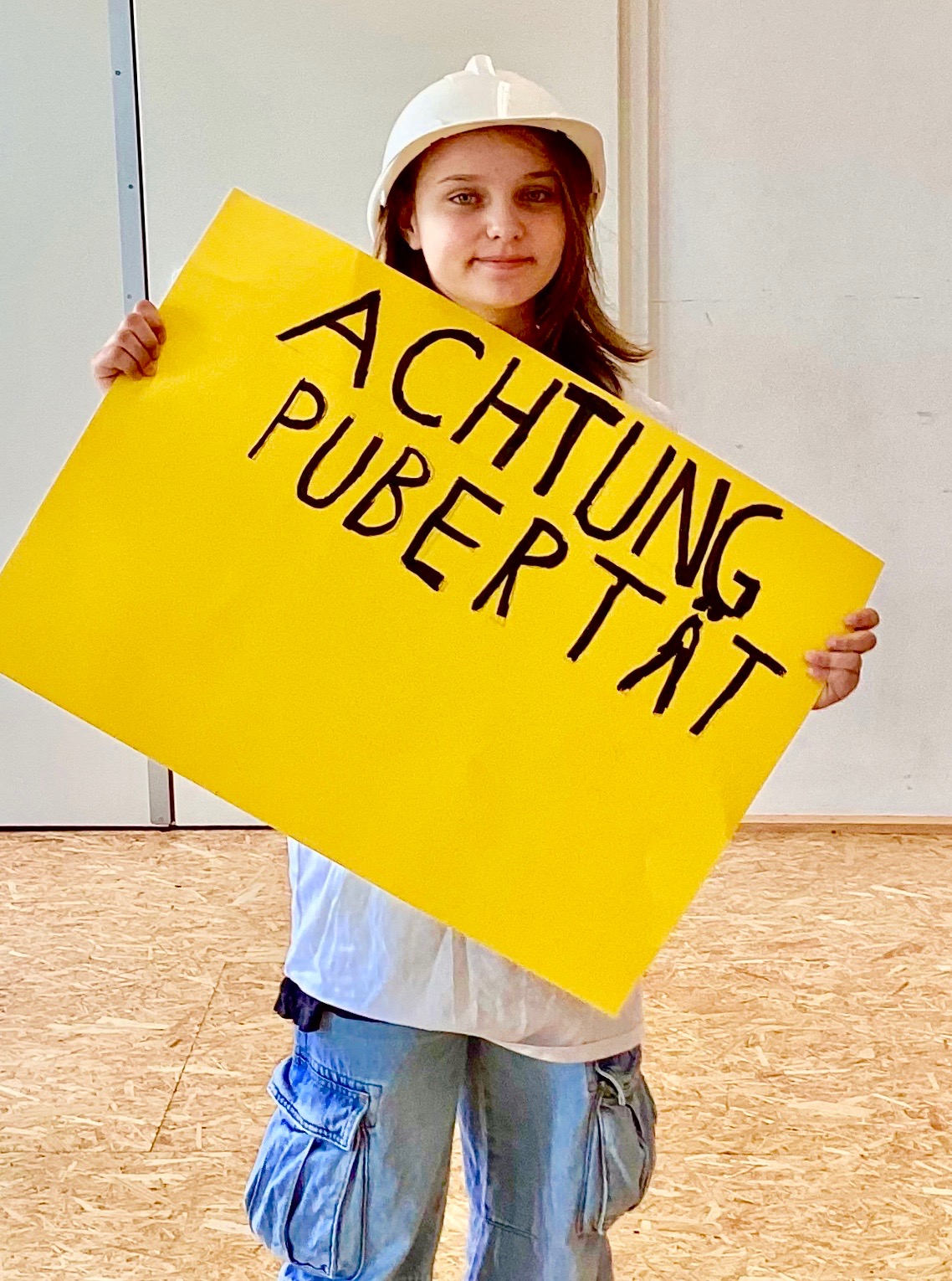 junges Mädchen mit Baustellenhelm hält ein großes gelbes Schild mit der Aufschrift "Pubertät"