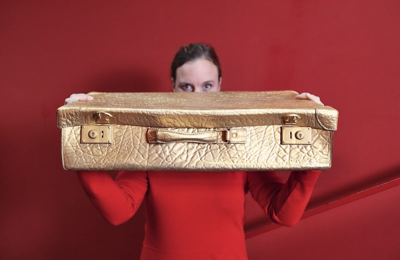 Kofferbühne. Theaterpädagogin Eva Ockelmann in rotem Kleid von rotem Hintergrund mit goldenem Koffer in der Hand