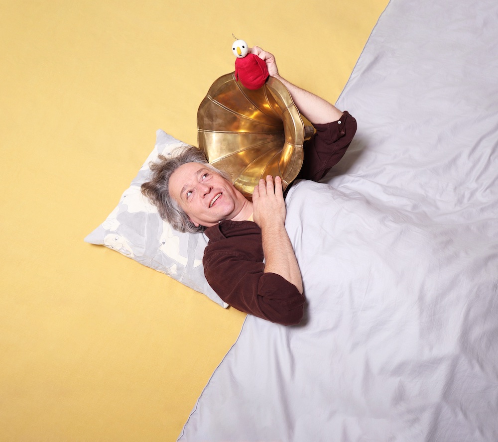 Weißnich. Christof Lappler unter grauer Decke mit dem Weißnich, einer kleinen Figur mit Augen, und einem goldenen Grammophon in der Hand.